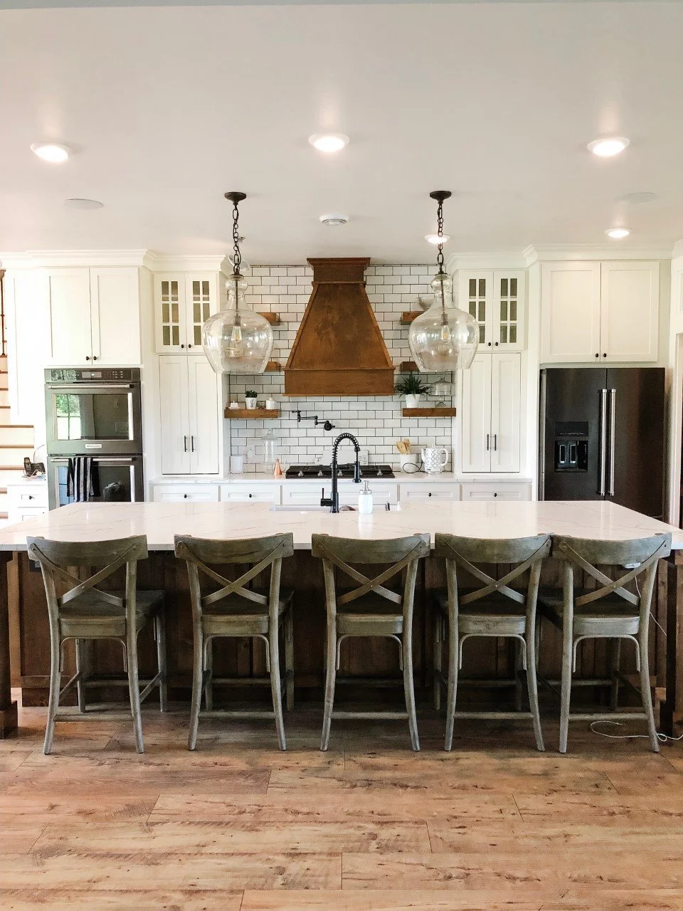 This beautifully remodeled kitchen features classic white shaker-style cabinets, a rustic wood range hood, and a large quartz-topped island with wood base cabinetry. Finished with subway tile backsplash, glass globe pendant lights, and wide-plank LVP flooring, this space blends farmhouse warmth with modern elegance. Cabinets by LaValle Flooring — proudly serving Valley City and Jamestown, ND.